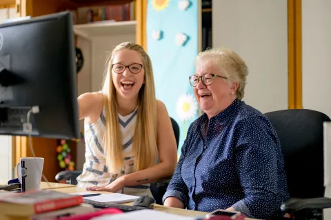 An older and a younger woman laughing and working together in front of a screen