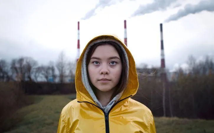 Image shows a photo of a young person in a yellow raincoat standing in front of 3 factory chimneys with light grey smoke coming from them.
