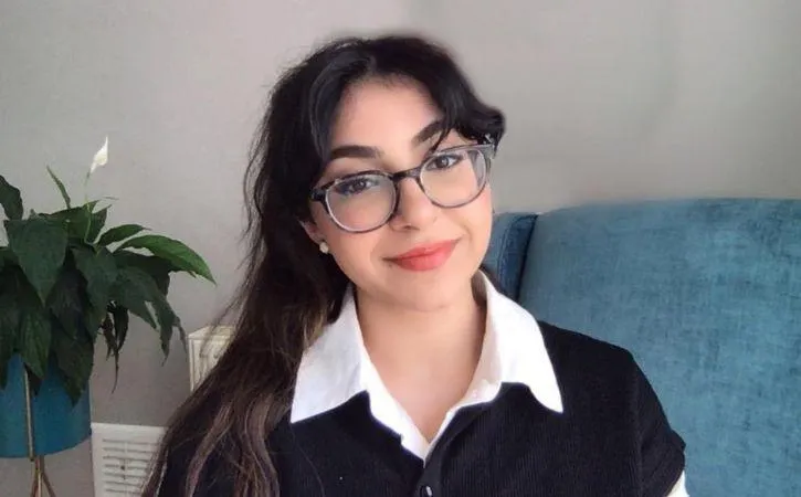 picture of a young person with long dark hair and glasses, sitting in front of a blue sofa and lily plant. The wall in the background is grey. 