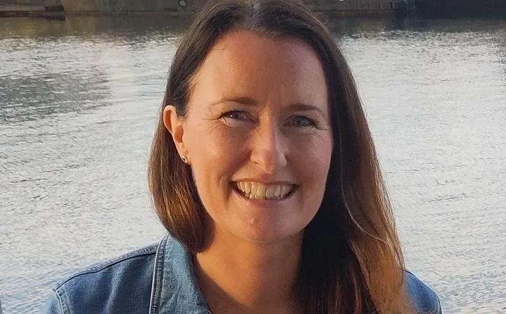Picture of a woman with brown hair looking straight into the camera and smiling, in front of a lake.