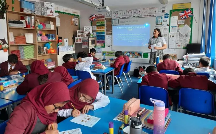 photo of a classroom of students sitting at blue desks, writing. There is a teacher at the front of the class, next to projected presentation on a white board. There are bookshelves around the room and colourful signs and art on the walls.
