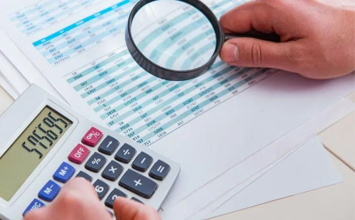 image shows a hand holding a magnifying glass over a spreadsheet with data. The other hand is using a calculator.