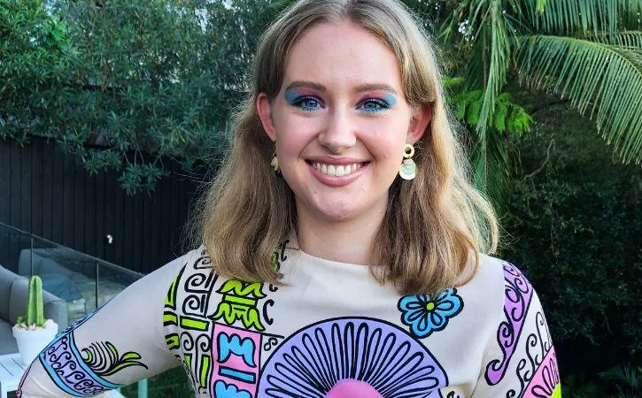 image shows a person with shoulder length blonde hair and colourful makeup, wearing a colourful shirt. The background is foliage. 