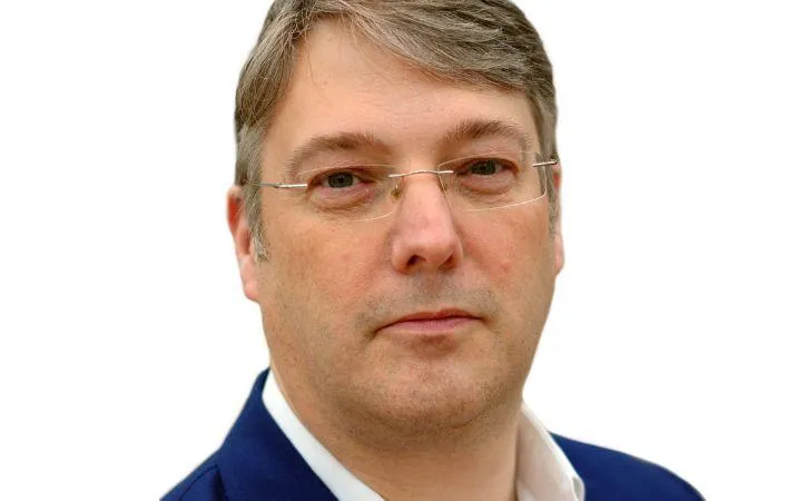 picture of a man with glasses looking straight into the camera with a navy jacket and white shirt.