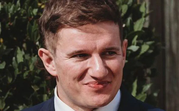 image shows a young man with light brown hair, wearing a white shirt and black jacket, smiling.