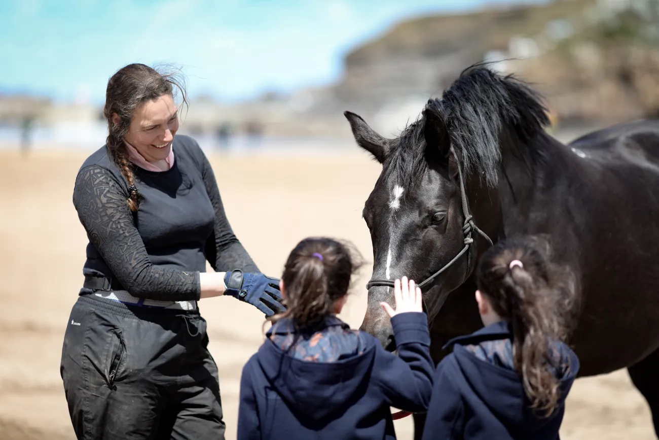 A smiling woman with long dark hair support two young children to greet a black horse with a white spot on its nose