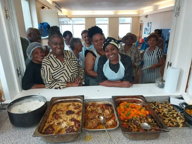 Image shows the products of a cookery class at the OK Foundation with trays of food in the foreground the class members standing behind and smilling.
