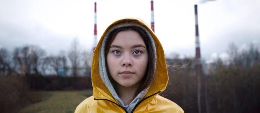 Image shows a photo of a young person in a yellow raincoat standing in front of 3 factory chimneys with light grey smoke coming from them.