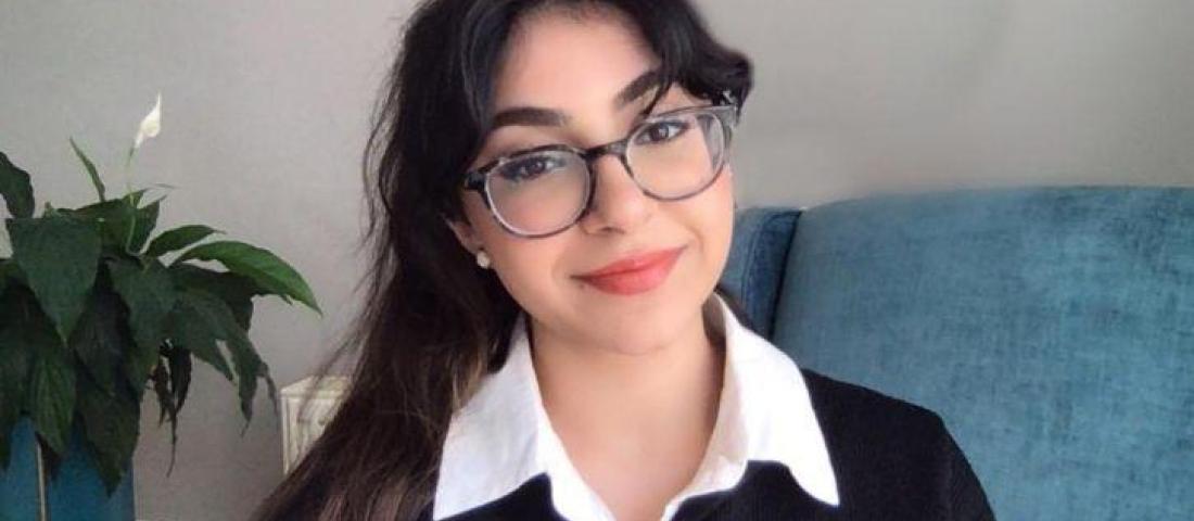 picture of a young person with long dark hair and glasses, sitting in front of a blue sofa and lily plant. The wall in the background is grey. 