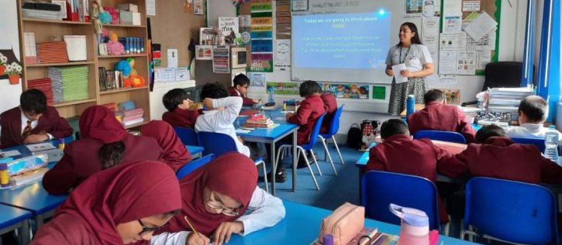 photo of a classroom of students sitting at blue desks, writing. There is a teacher at the front of the class, next to projected presentation on a white board. There are bookshelves around the room and colourful signs and art on the walls.