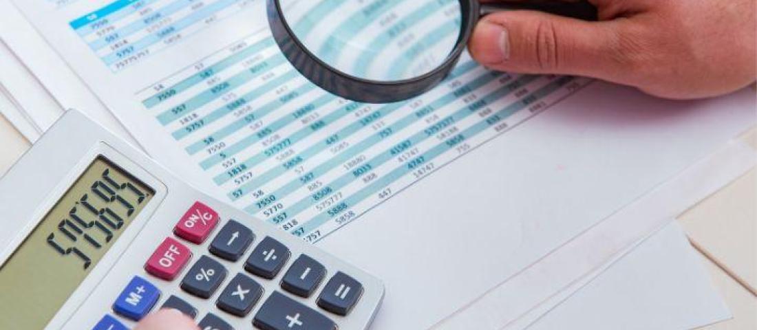 image shows a hand holding a magnifying glass over a spreadsheet with data. The other hand is using a calculator.
