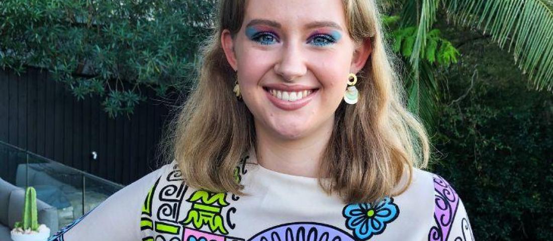image shows a person with shoulder length blonde hair and colourful makeup, wearing a colourful shirt. The background is foliage. 