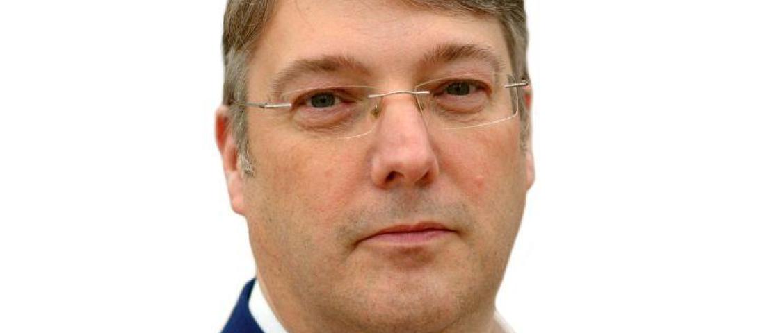 picture of a man with glasses looking straight into the camera with a navy jacket and white shirt.