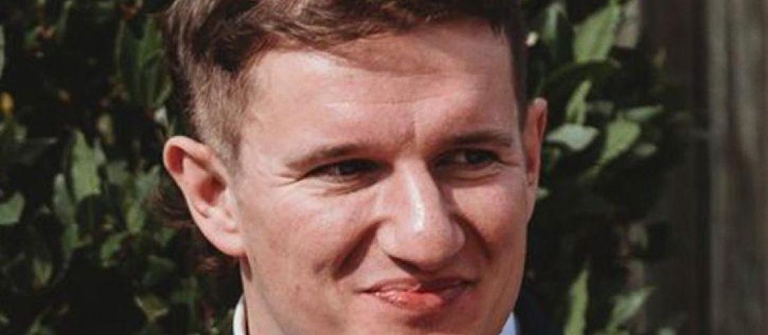 image shows a young man with light brown hair, wearing a white shirt and black jacket, smiling.