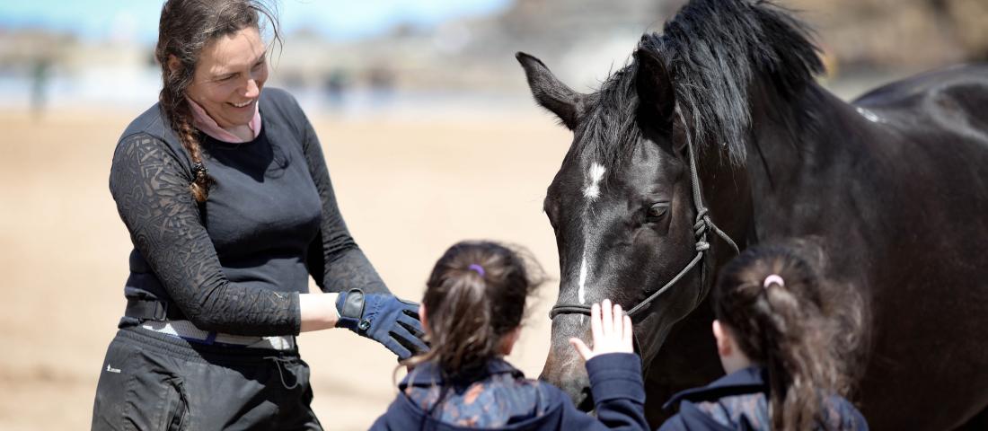A smiling woman with long dark hair support two young children to greet a black horse with a white spot on its nose