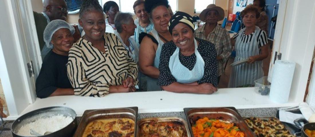 Image shows the products of a cookery class at the OK Foundation with trays of food in the foreground the class members standing behind and smilling.