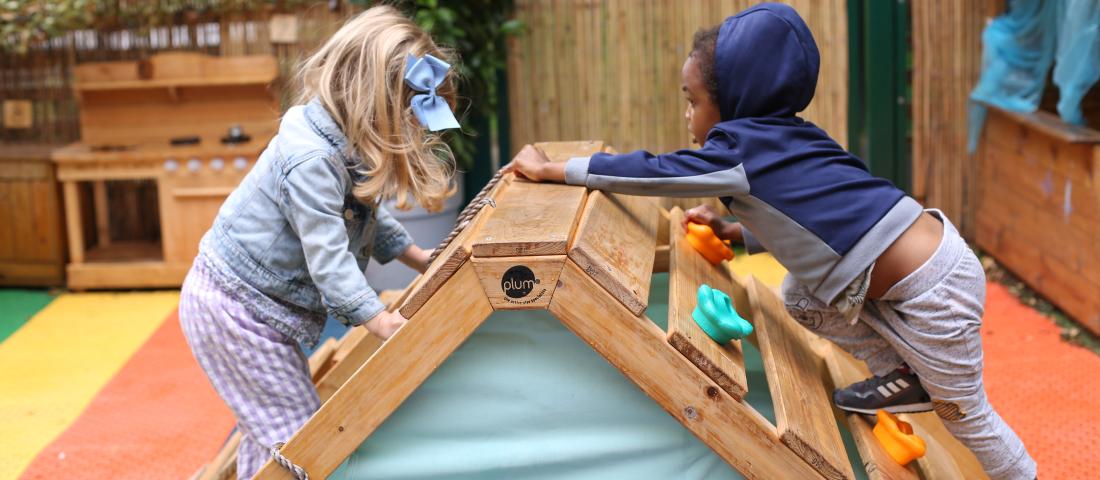 Two young children playing on a low wooden climbing wall. 